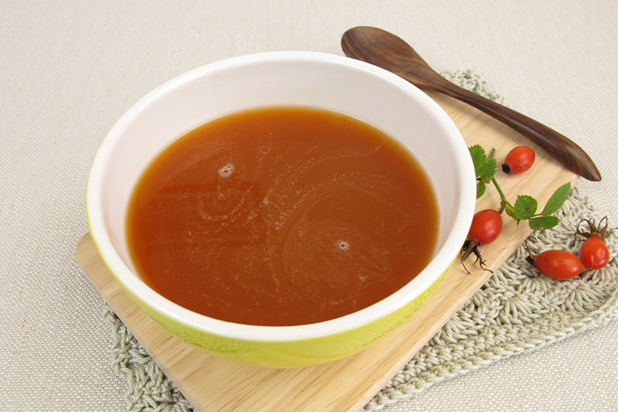 Cremig rötliche Suppe in gelber Schüssel auf Holzbrett. Daneben ein Holzlöffel und einige Hagebutten mit Blättern.